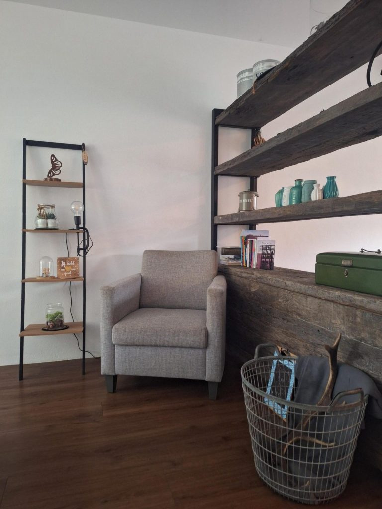 Welkom in een warme en rustige ruimte voor verbinding en groei. Coach en therapeut. Een comfortabele grijze stoel naast een open boekenkast en een mand op de vloer. Coach en therapeut.