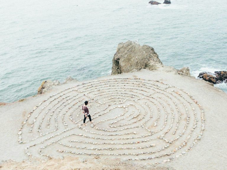 Stap voor stap dichter bij jezelf. Coach en therapeut. Een persoon wandelt door een zandlabyrint aan de kust. Coach en therapeut.